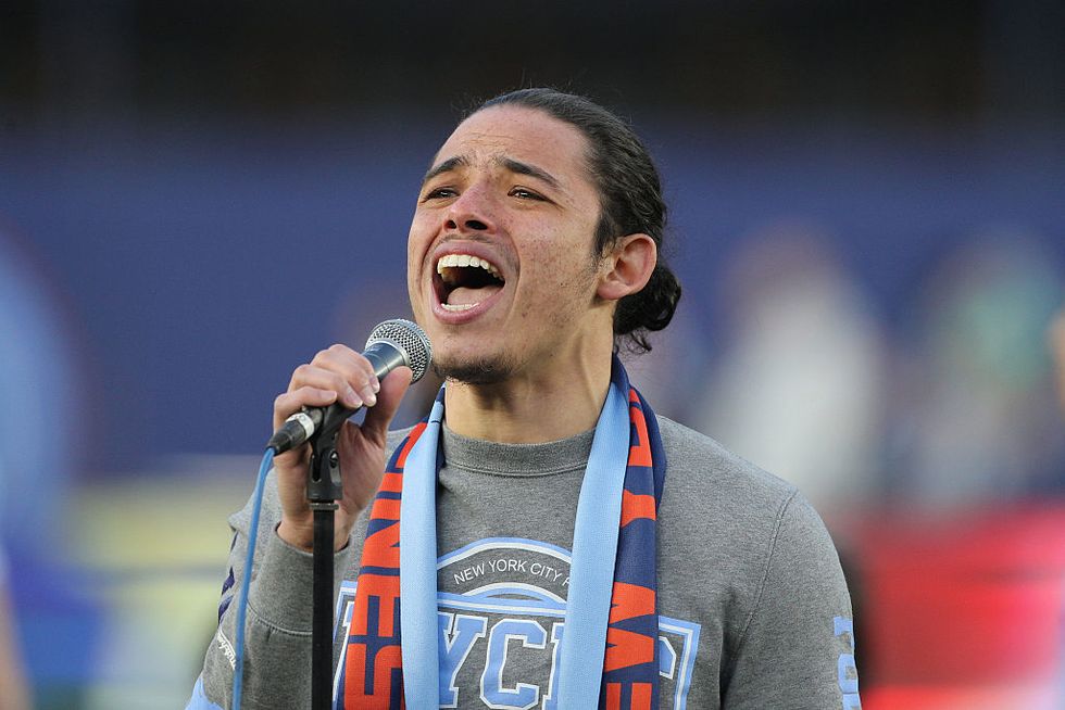 anthony ramos, wearing a gray t shirt and blue scarf, sings into a microphone while standing outside