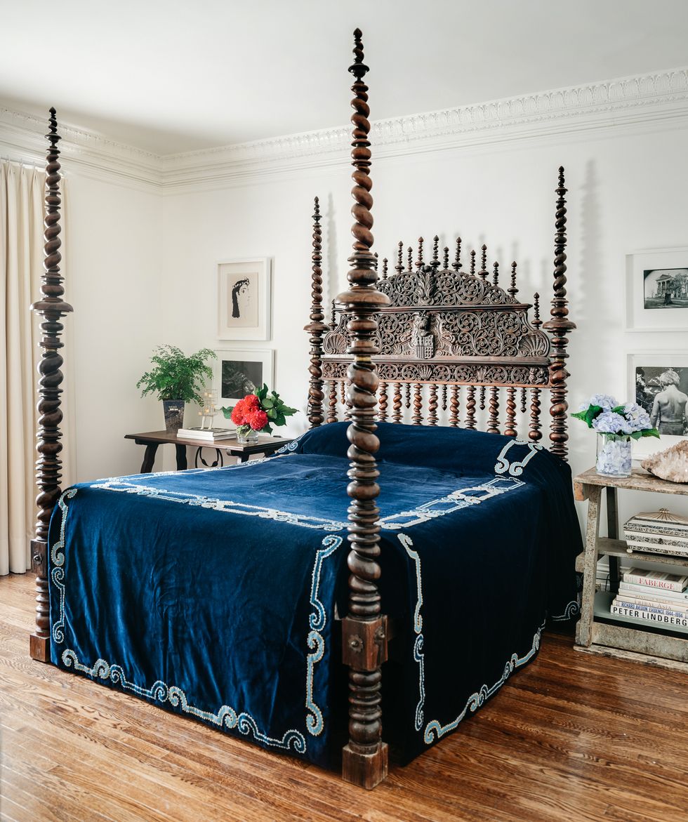 antique four poster bed with spirally posts and headboard and a heavy dark blue velvet cover with with stitching and two worn wood night stands that look like sawhorses and some framed prints above them