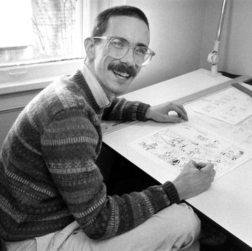 bill watterson, wearing glasses and a mustache, smiles at the camera while sitting at a desk and drawing a cartoon