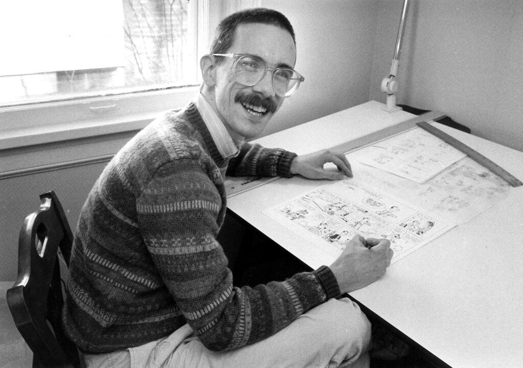 bill watterson, wearing glasses and a mustache, smiles at the camera while sitting at a desk and drawing a cartoon