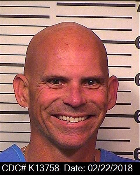 lyle menendez smiles in a mug shot, he wears a blue prison shirt and a chain around his neck