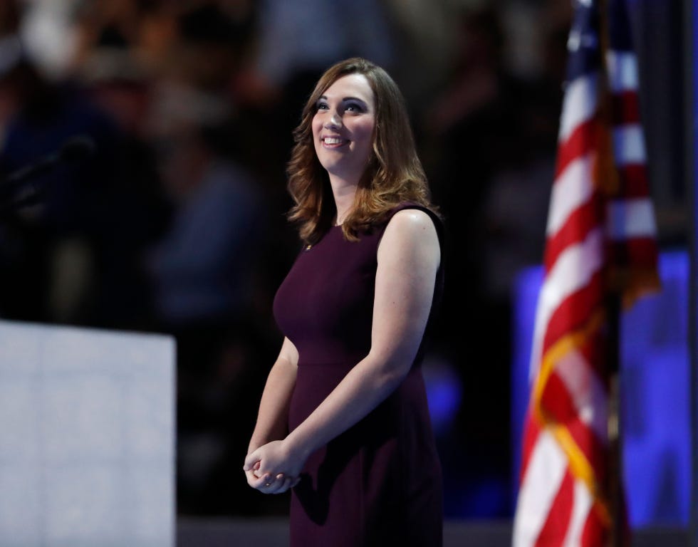 sarah mcbride smiles and looks up as she stands with her hands clasped on a stage, she wears a purple sleeveless dress, an american flag is behind her on the right