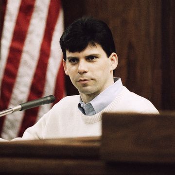 lyle menendez sits on the witness stand in a wood paneled room, he wears a white sweater and a blue collared shirt, behind him is an american flag and a white bulletin board with a blurry photo of a woman