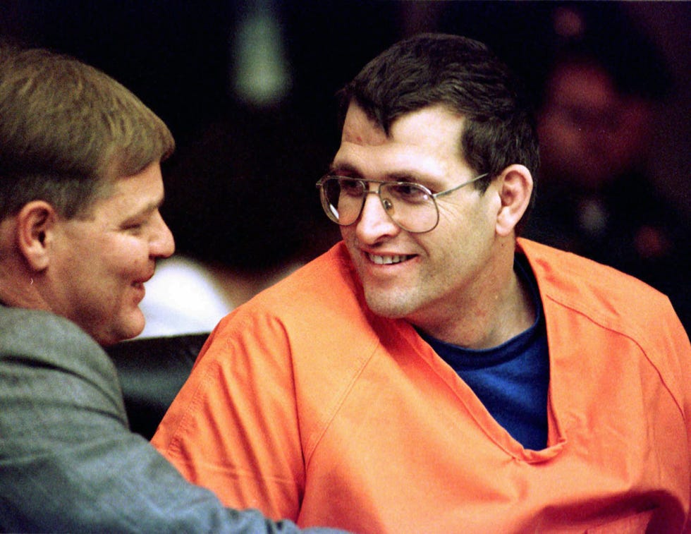 a man in an orange shirt and glasses talking with another person in suit