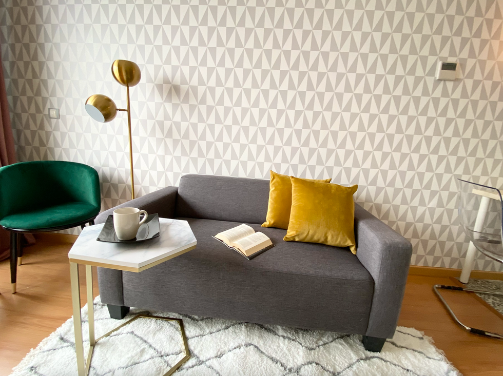 salón moderno con sofá gris, mesa auxiliar de mármol, papel pintado geométrico y butaca de terciopelo verde con lámpara de pie dorada
