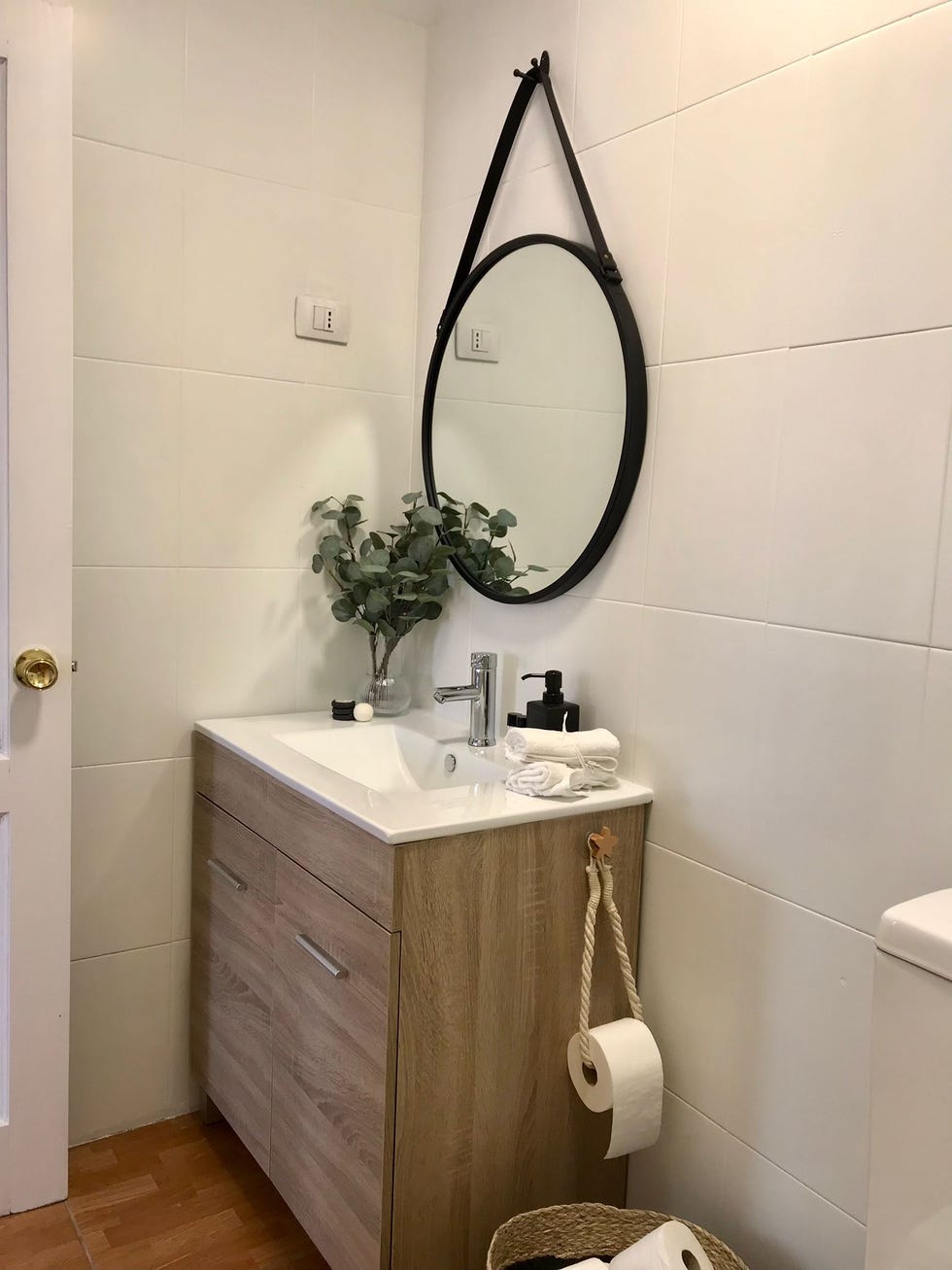 baño con mueble de madera y espejo redondo con marco metálico negro y correa