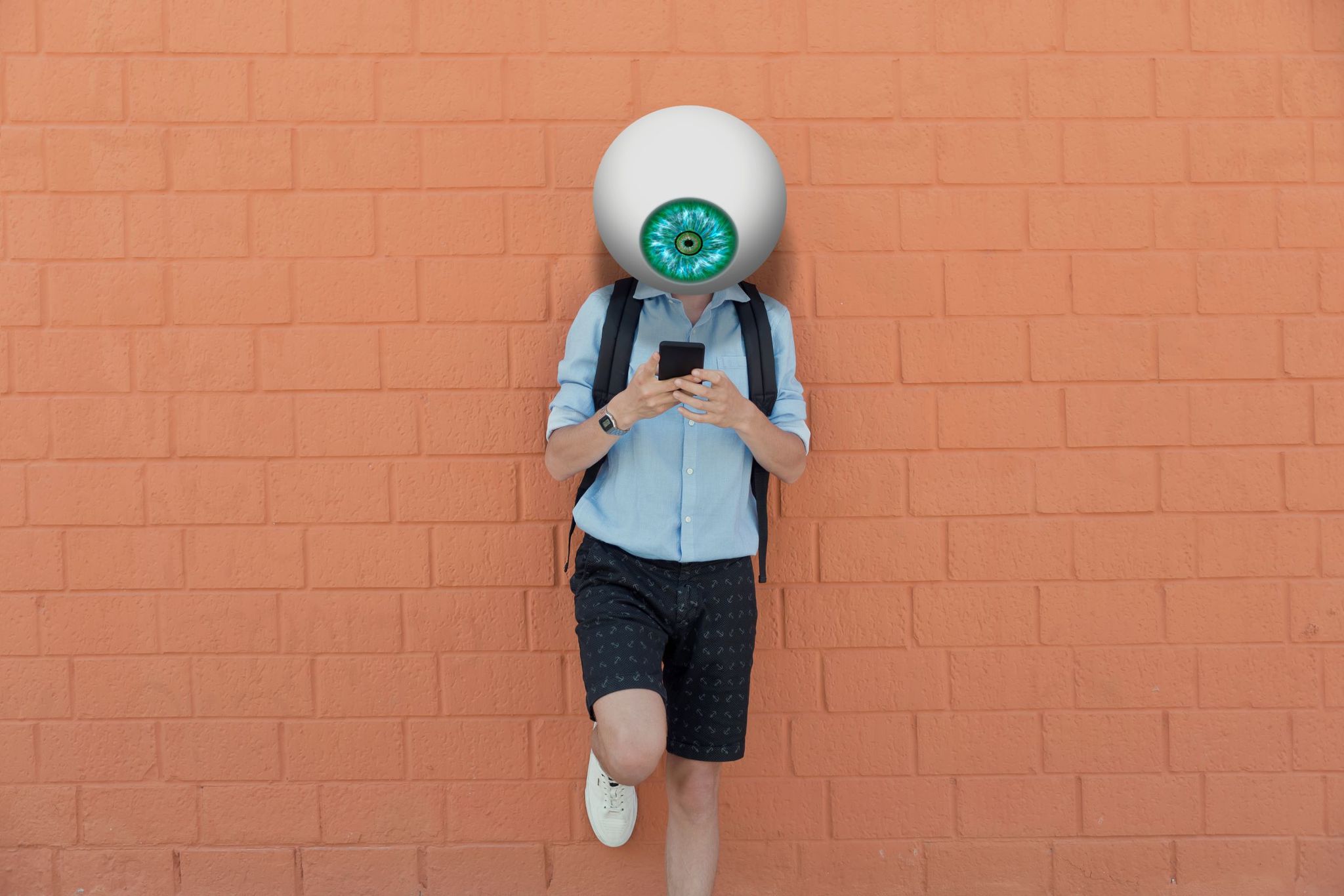 un enorme ojo artificial sustituye la cabeza de un joven con camisa azul y pantalón corto de cuadros oscuro que está consultando su teléfono móvil, apoyado en un pared