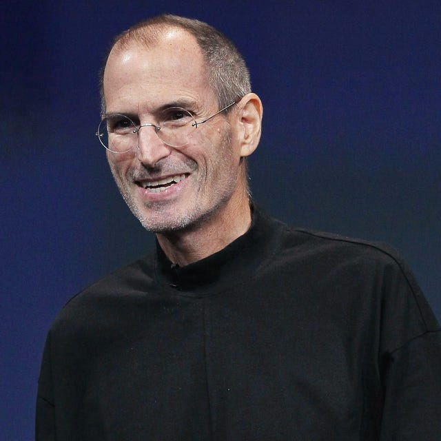 steve jobs smiles and looks past the camera, he is wearing a signature black turtleneck and circular glasses with a subtle silver frame, behind him is a dark blue screen