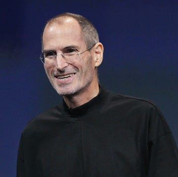 steve jobs smiles and looks past the camera, he is wearing a signature black turtleneck and circular glasses with a subtle silver frame, behind him is a dark blue screen