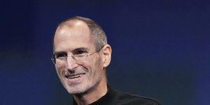steve jobs smiles and looks past the camera, he is wearing a signature black turtleneck and circular glasses with a subtle silver frame, behind him is a dark blue screen