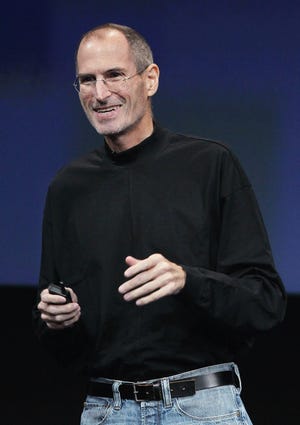 steve jobs smiles and looks past the camera, he is wearing a signature black turtleneck and circular glasses with a subtle silver frame, behind him is a dark blue screen