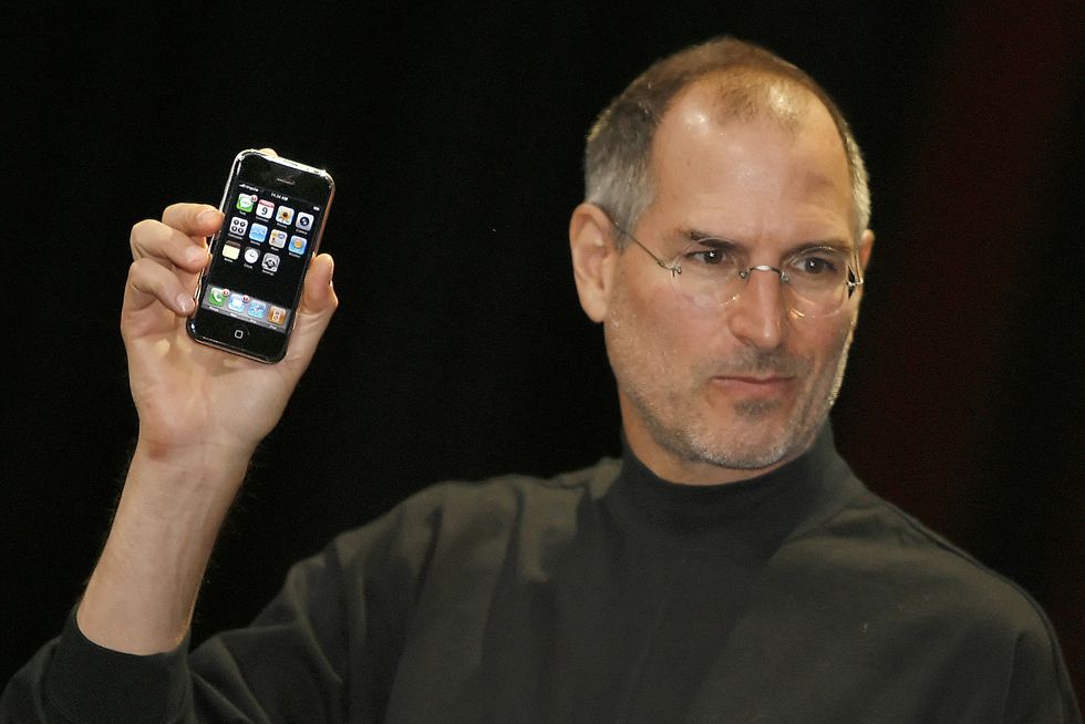 steve jobs, wearing a black long sleeved shirt and glasses, holding up an iphone and looking off camera