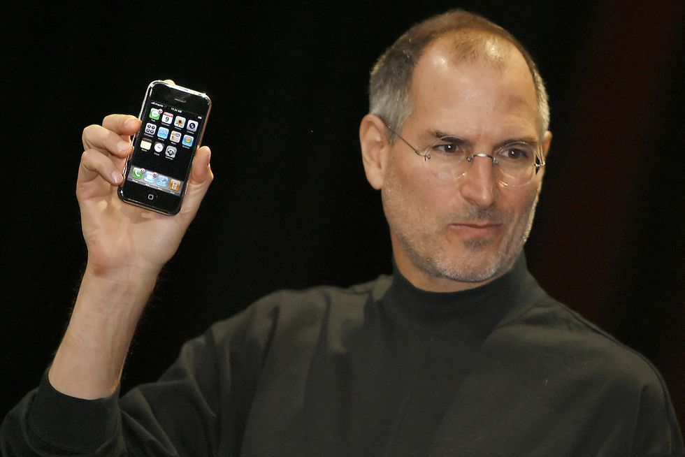 steve jobs, wearing a black long sleeved shirt and glasses, holding up an iphone and looking off camera