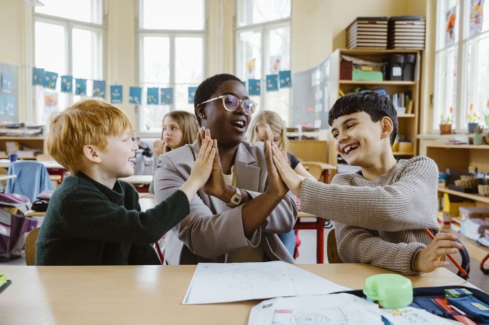 happy teacher and boys giving high fives while sitting at desk in classroom