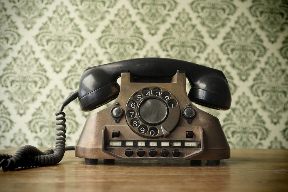 old telephone made of copper resting on table, vintage green patterned wallpaper on wall behind it