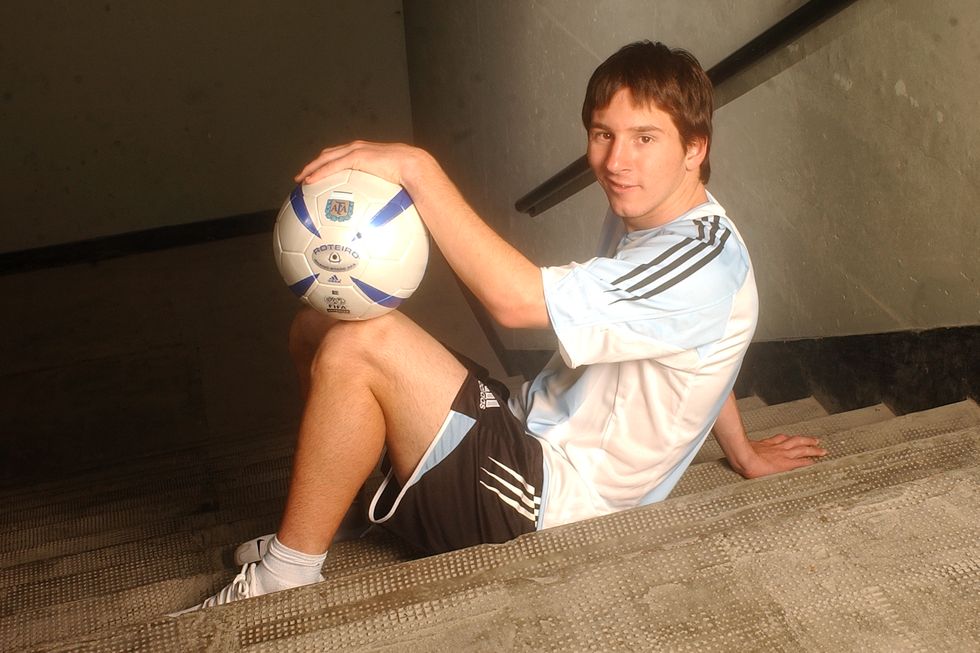 lionel messi sits in a stairwell and holds a soccer ball on his knees, he smiles at the camera and wears an argentina national team uniform