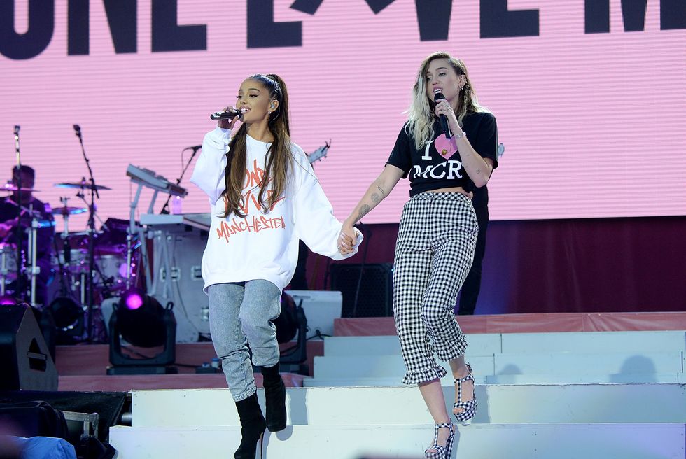 ariana grande and miley cyrus walk down stairs on a stage holding hands, both sing into wireless microphones