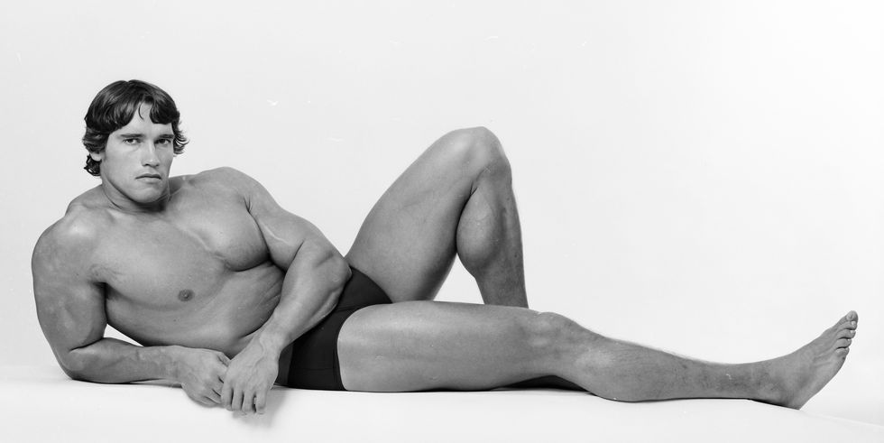 arnold schwarzenegger lies down with one leg extended and the other with his knee bent and foot on the ground, he wears short shorts and no other clothing and looks at the camera with a seductive look on his face
