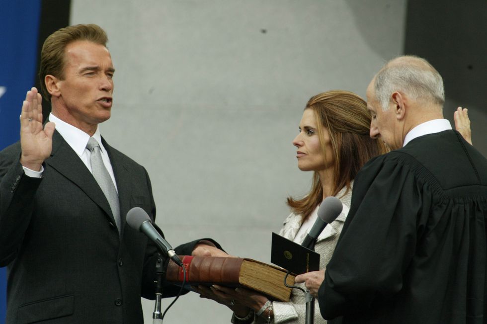 arnold schwarzenegger has one hand raised in the air and another resting on a book held by wife maria shriver during his swearing in ceremony to become california governor, the judge performing the ceremony is on the right
