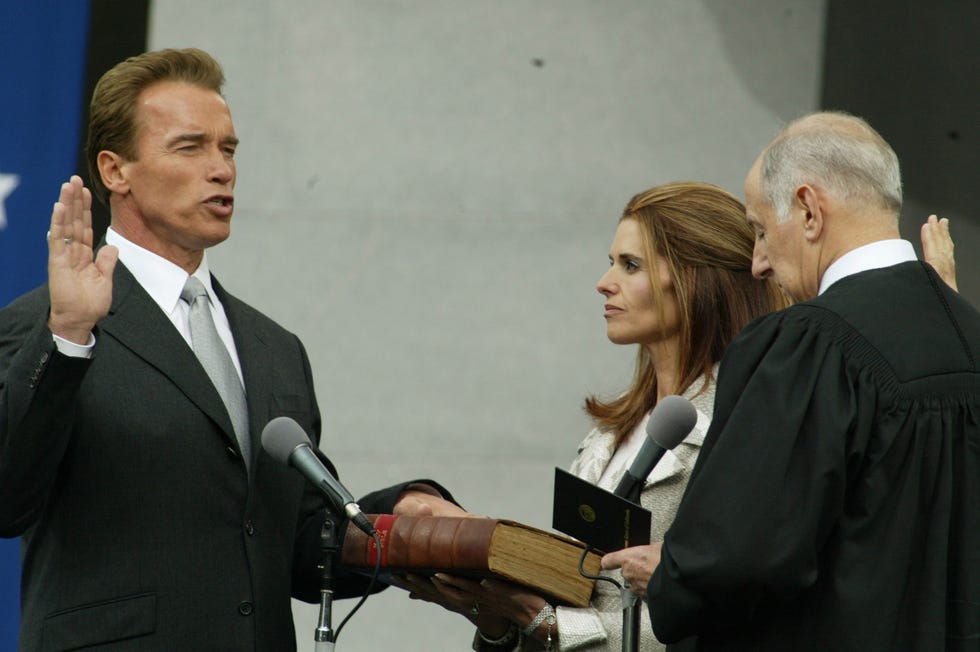 arnold schwarzenegger has one hand raised in the air and another resting on a book held by wife maria shriver during his swearing in ceremony to become california governor, the judge performing the ceremony is on the right