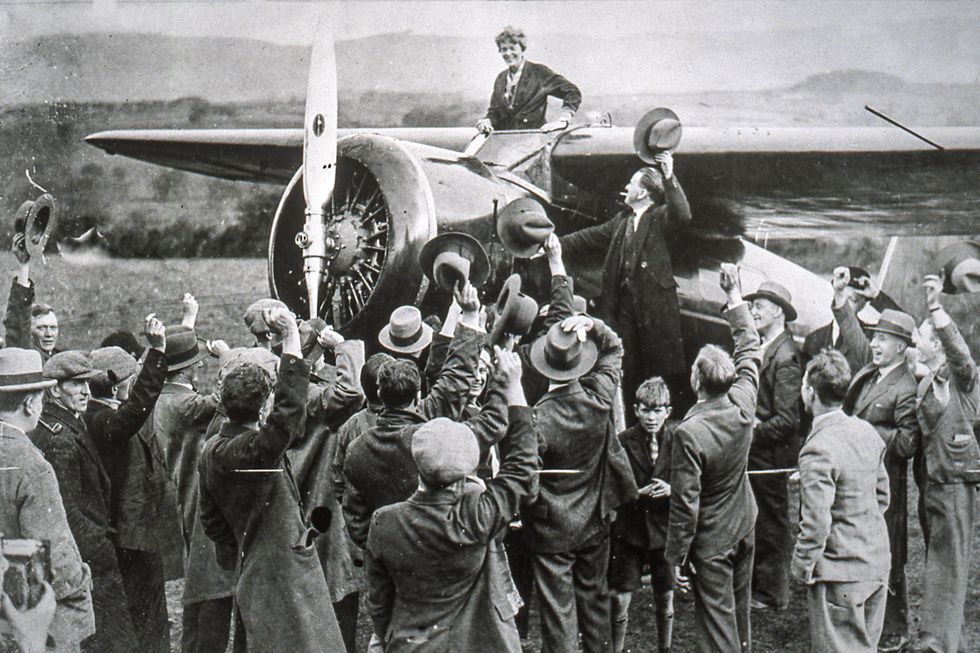 amelia earhart stands in a plane cockpit and smiles down at a crowd of people next to the plane, many people wave and lift hats to her