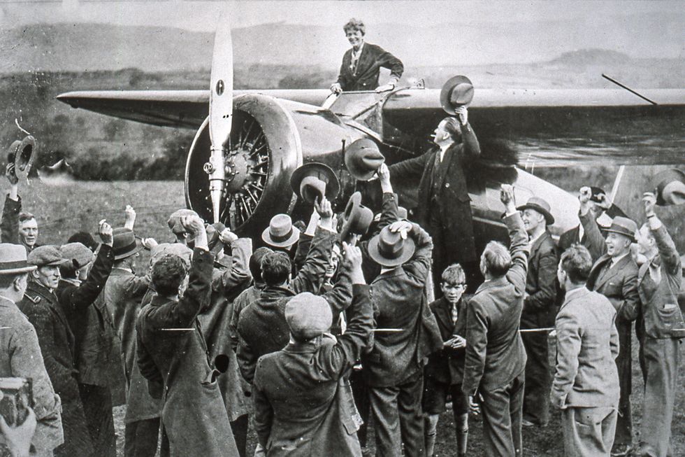 amelia earhart stands in a plane cockpit and smiles down at a crowd of people next to the plane, many people wave and lift hats to her