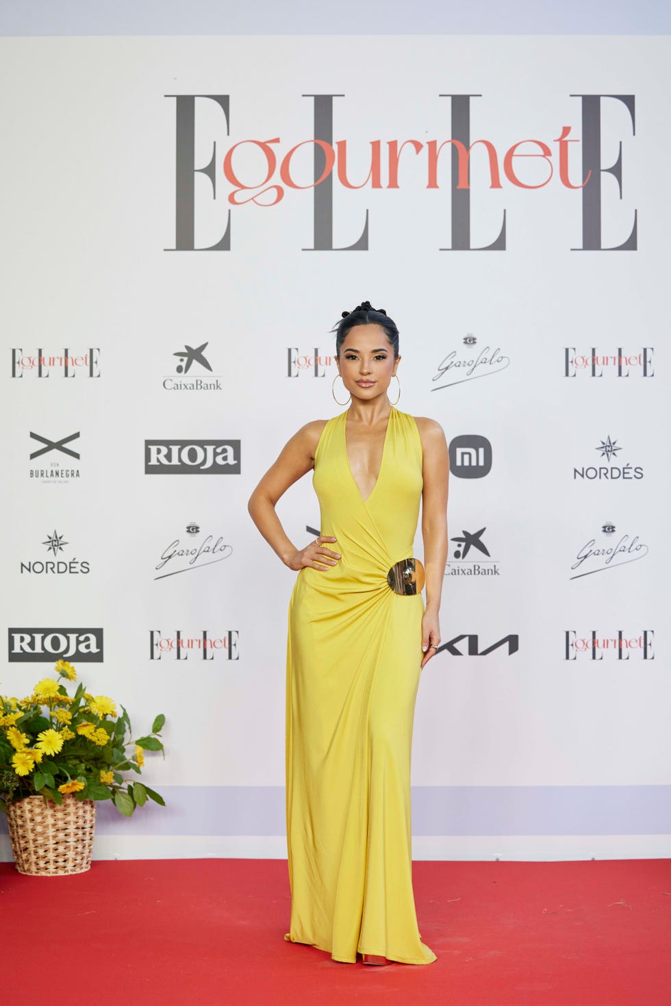 A woman wearing a yellow gown poses on a red carpet at an event