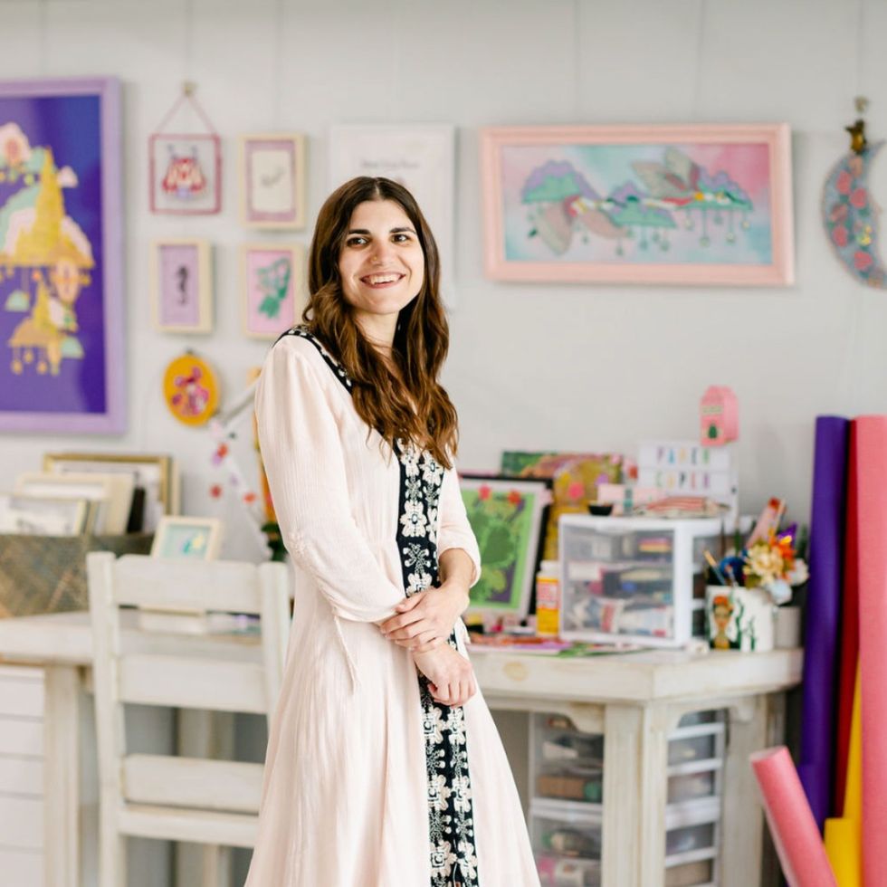 woman standing in front of a desk with paintings and craft supplies around her