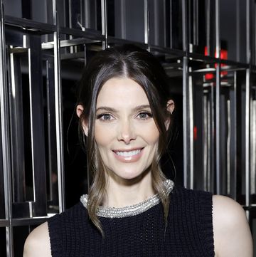 ashley greene, a young woman stands with her left hand in trouser pocket, smiling at the camera, she has brown hair worn back and wears a black top and trousers