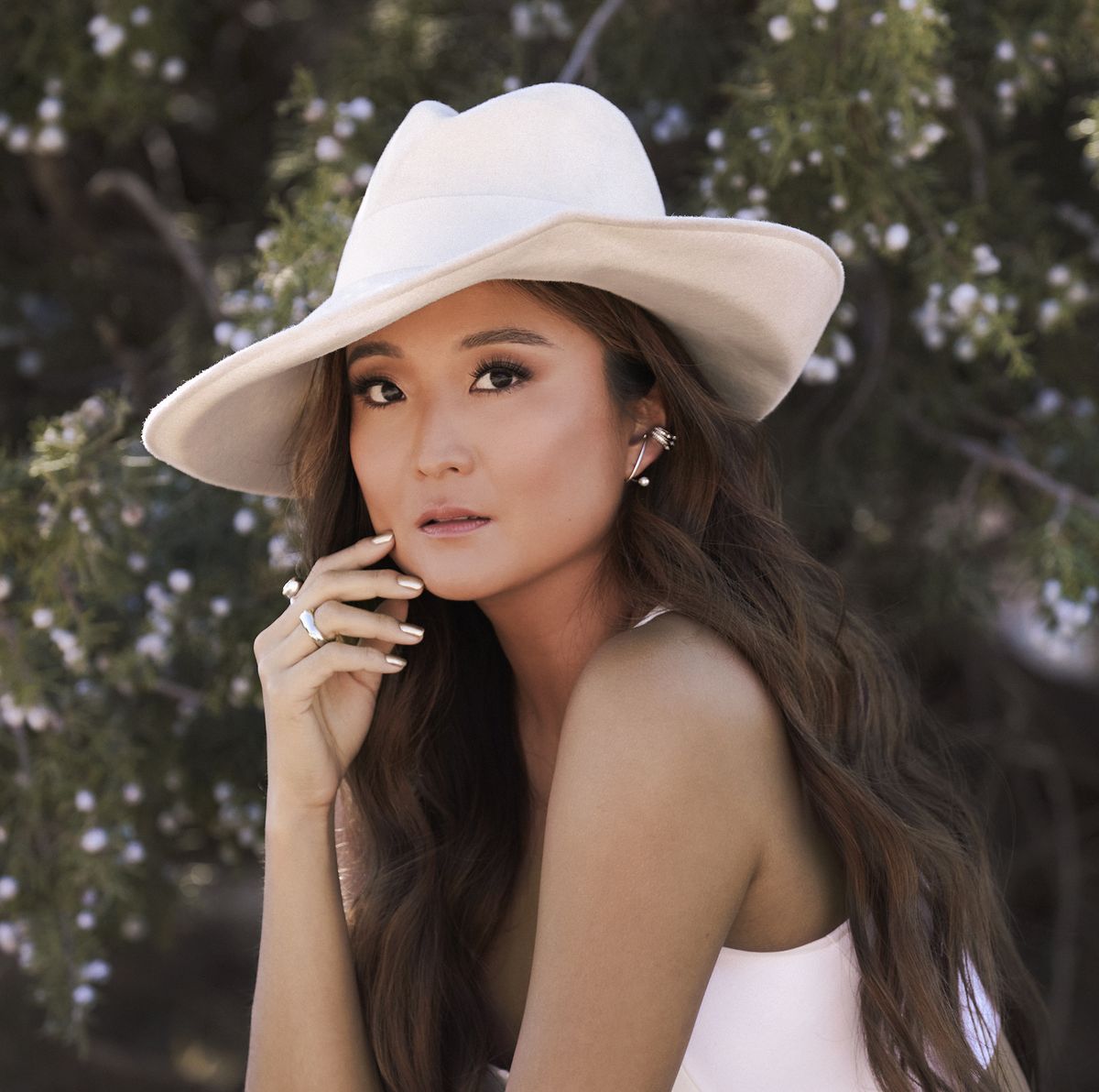 ashley park poses in front of plants wearing cream fedora hat, white tank top, and jewelry