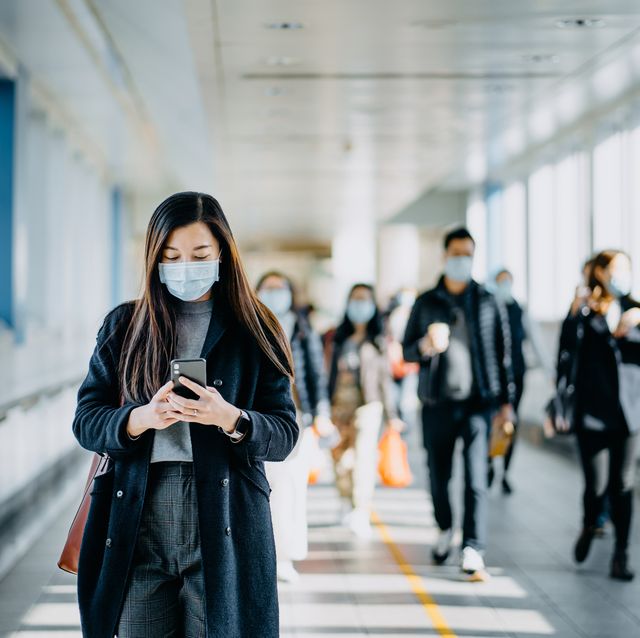 asian woman with protective face mask using smartphone while commuting in the urban bridge in city against crowd of people