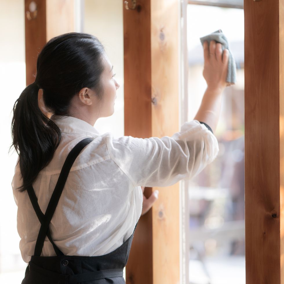asian young woman wiping the window glass of a cafe restaurant wearing an apron with a ponytail