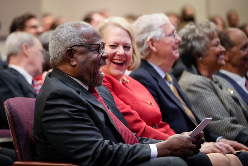 clarence thomas, wearing a black suit and red tie, laughs along with his wife, virginia thomas, who are both seated in a crowded room