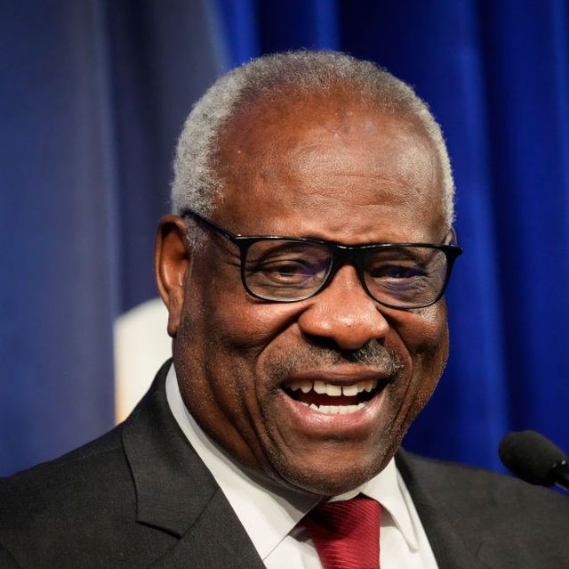 supreme court justice clarence thomas smiling and standing in front of a microphone, he is wearing a black suit jacket, white collared shirt, red tie, and black framed glasses