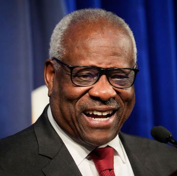 supreme court justice clarence thomas smiling and standing in front of a microphone, he is wearing a black suit jacket, white collared shirt, red tie, and black framed glasses