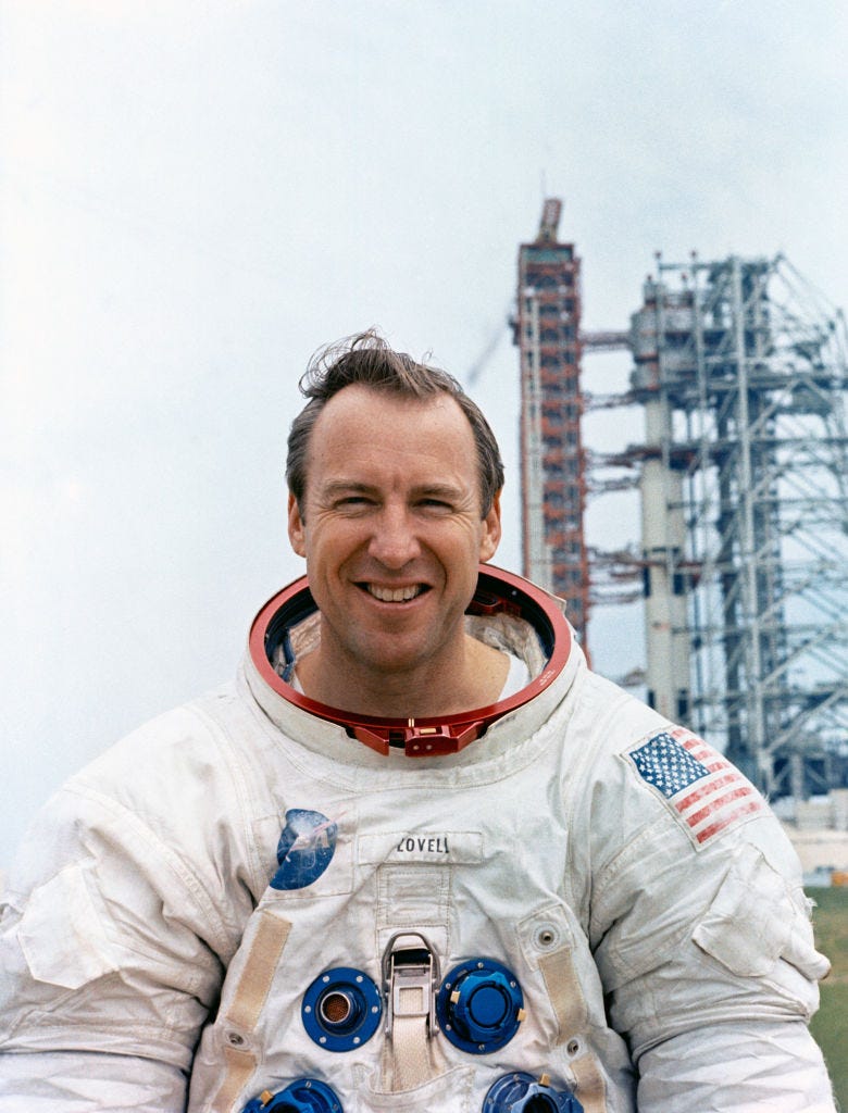 Jim Lovell wears his spacesuit and smiles in front of a large Space Shuttle Dock