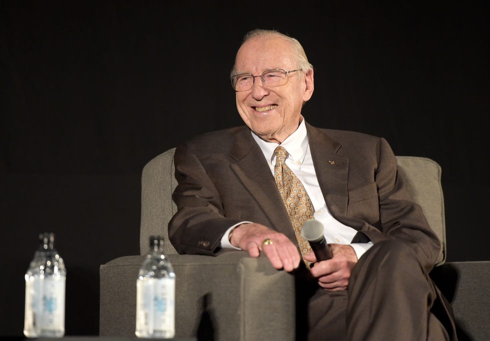 Astronaut Jim Lovell wears a suit and sits on a chair during a panel discussion