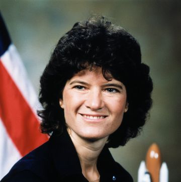 a woman smiles as she looks left, she wears a dark collared top, part of an american flag and a model space shuttle are in the background