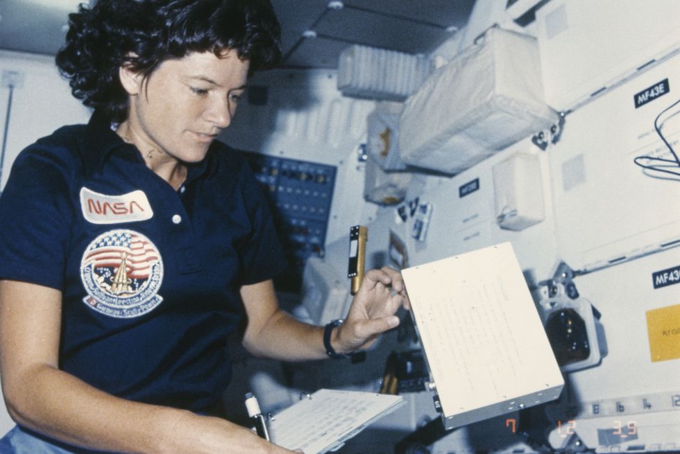 a woman in a nasa uniform holds a pen and paper as she reaches for a floating object in front of her