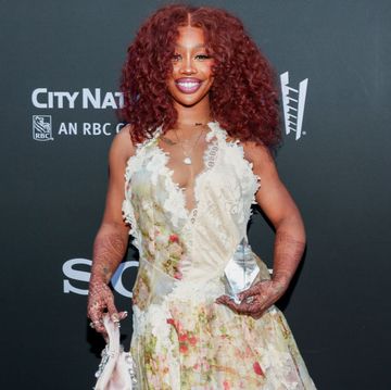 sza smiles at the camera while standing in front of a gray background, she wears a lace and floral dress and holds a crystal award in one hand by her waist and a purse in the other hand