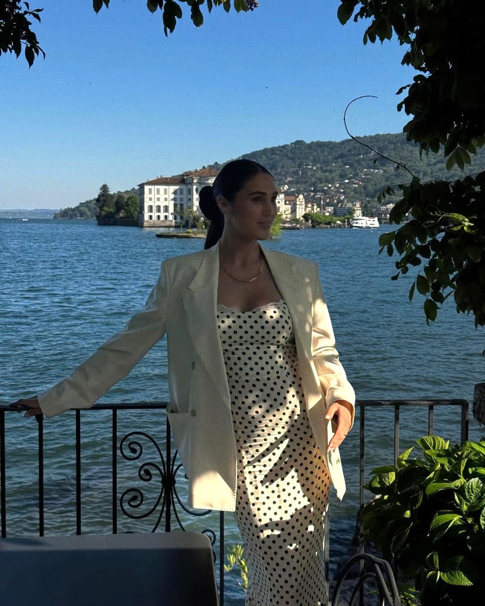 a woman in a polka dot dress and blazer poses by a scenic waterfront