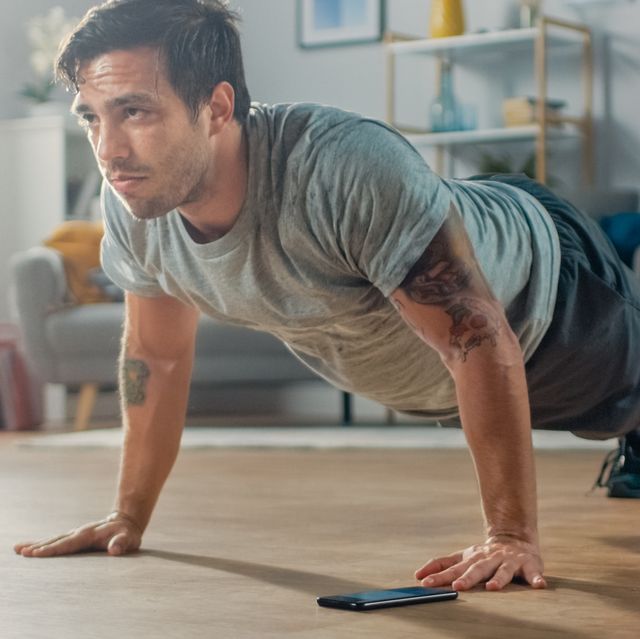 athletic fit man in t shirt and shorts is doing push up exercises while using a stopwatch on his phone he is training at home in his living room with minimalistic interior