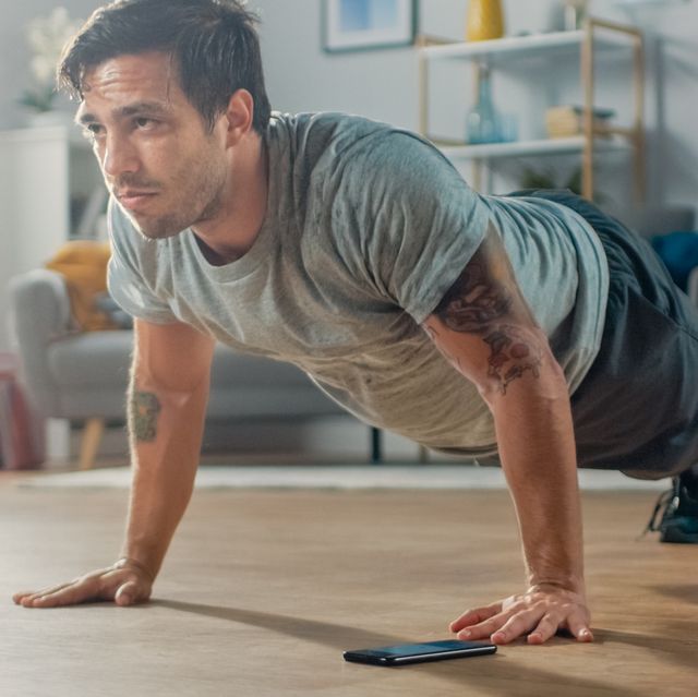 athletic fit man in t shirt and shorts is doing push up exercises while using a stopwatch on his phone he is training at home in his living room with minimalistic interior