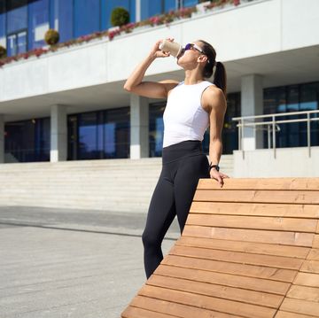 athletic woman in activewear drinking a smoothie outdoors, leaning on a wooden bench after workout in a modern urban area.