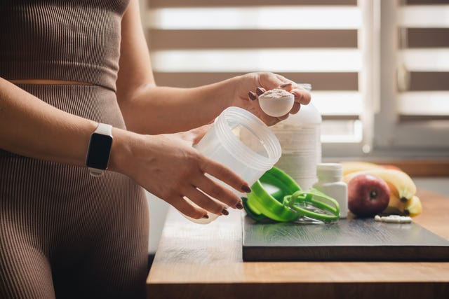 athletic woman in sportswear with measuring spoon in her hand puts portion of whey protein powder into a shaker on wooden table with amino acid white capsules, bananas and apple, making protein drink