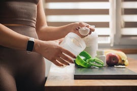 athletic woman in sportswear with measuring spoon in her hand puts portion of whey protein powder into a shaker on wooden table with amino acid white capsules, bananas and apple, making protein drink