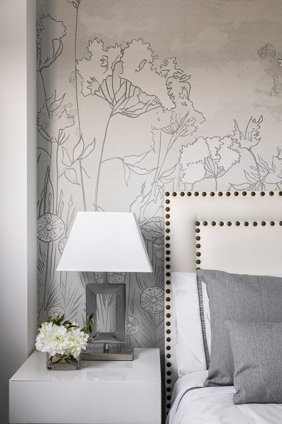 dormitorio con cabecero acolchado en blanco con remaches y papel pintado con motivos florales en gris
