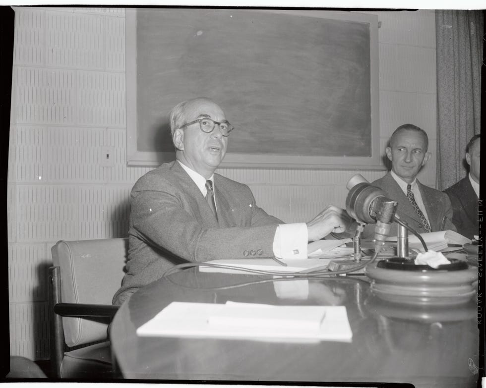 lewis l strauss sits and talks at a table with papers in front of him and a chalkboard on the wall behind