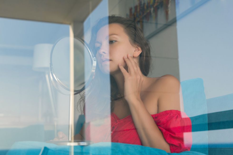 Attractive young woman looking at mirrow behind the glass of window her room.