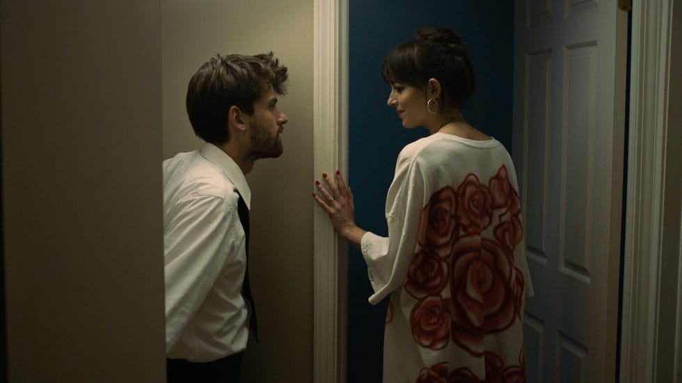 In a narrow hallway, a man wearing a white shirt and black tie leans toward a closed door, showing curiosity or concern. He stands near the doorframe and faces a woman standing next to him